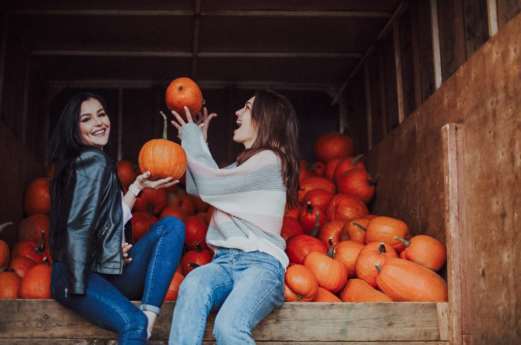 two girls smiling while holding pumpkins with bunch of pumpkins behind them